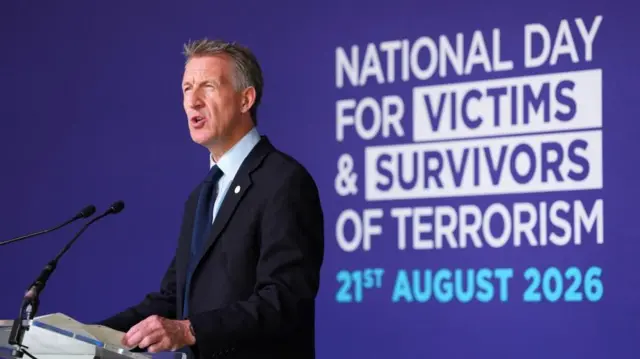 Dan Jarvis is pictured speaking from a podium with the words 'National Day For Victims and Survivors of Terrorism 21st August 2026' on a purple background behind him.