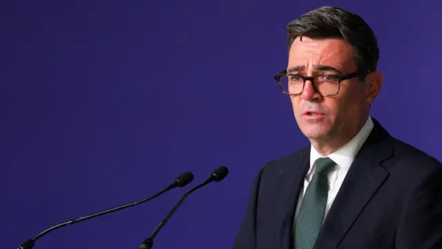 Andy Burnham is pictured speaking behind microphones against a purple background