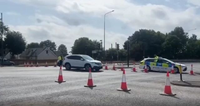 Two vehicles including a police car are shown blocking access to a major road, with traffic cones placed around them.