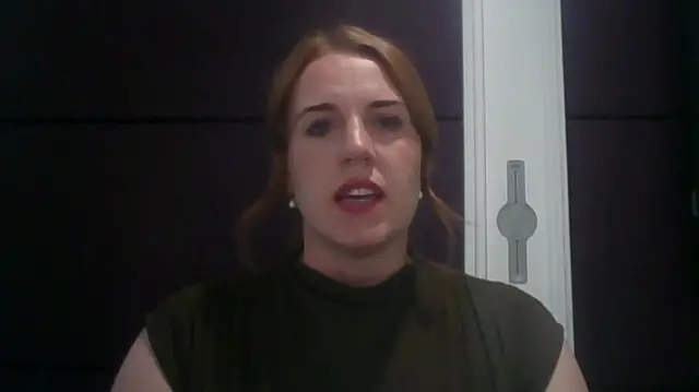 A woman wearing black speaking in front of a camera in a room