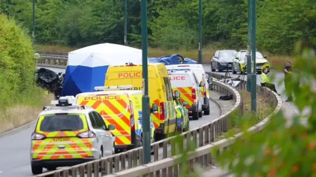 A number of emergency vehicles are shown at the scene of the crash, around a large blue and white tent on the motorway.