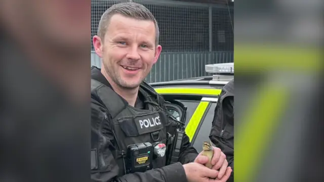 PC Tom Clough is pictured in police uniform, smiling and holding a duckling.