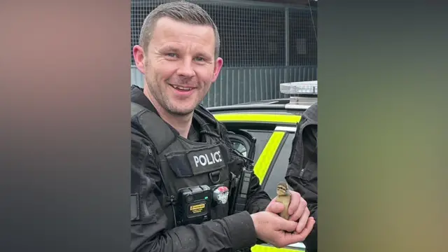 PC Tom Clough is wearing a black police uniform and a body cam. He is holding a baby duckling and smiling brightly at the camera. He has short brown hair and stubble.