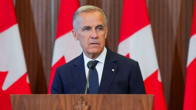 Mark Carney stands at a lecturn in a blue suit and tie with a white shirt