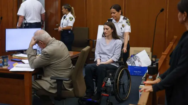 a view inside the courtroom showing an officer wheeling Clancy in on her wheelchair, beside her defense attorney with other officers in the background