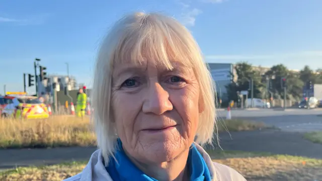 Sue Jeffrey is looking at the camera with a slightly sad expression on her face. She has mid-length white hair and is wearing a pale coat and blue scarf. There is an emergency vehicle and worker on the road behind her. Plus a collection of floral tributes under the traffic lights on the right.