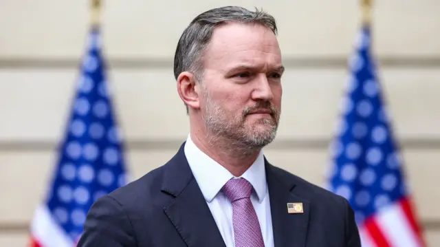 A man wears a suit with a pink tie and a US flag badge on his right lapel. He is looking off to the right side of the camera.