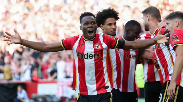 Brentford midfielder Mamadou Sangare celebrating a goal surrounded by his teammates. He is wearing a red-and-white Brentford home shirt.