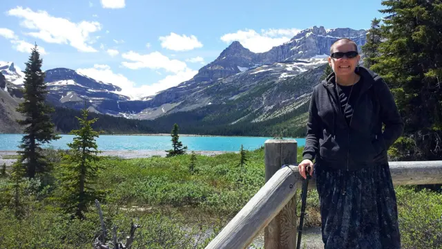 Cindy Baldassi is in the foreground wearing sunglasses and a dark hoody and navy blue dress. She is looking at the camera and behind her is a beautiful blue lake and towering mountain peaks with snow on top of them.