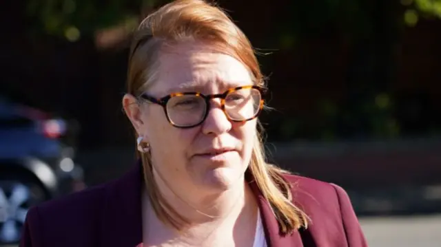 MP Anna Turley is standing in the street wearing burgundy blazer, white top and tortoiseshell glasses. She has ginger, shoulder length hair and is talking to someone off camera.