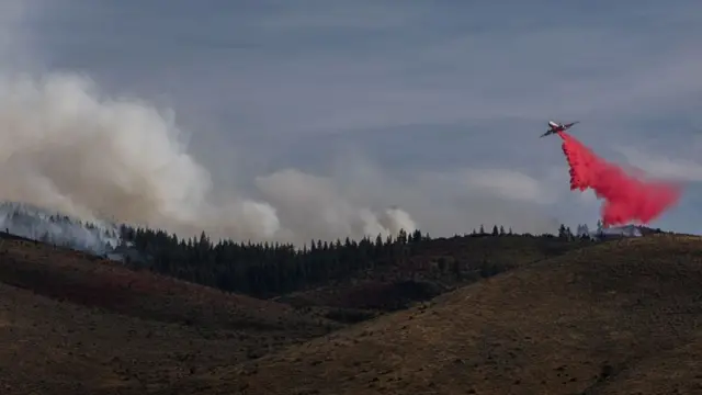 An aircraft flies over dry land that is on fire, as a large red cloud is visible beneath the plane as it drops fire retardant.