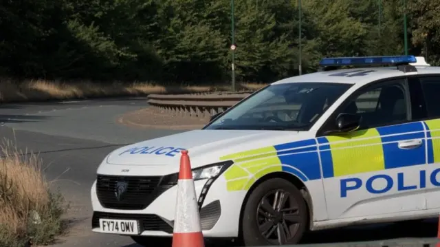 A police car is shown parked across a road, with an orange road cone in front of it.