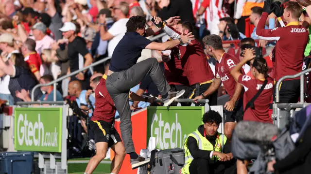 Brentford head coach Keith Andrews jumping into the dugout to celebrate a goal with his coaching staff. He is wearing a black shirt.