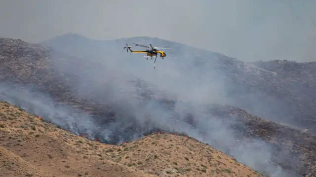 A helicopter flying over burning hillsides with smoke and fire rising from them