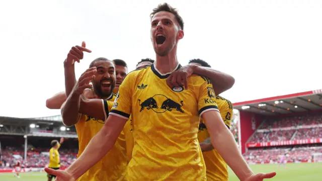 Anton Stach celebrating a late goal for Leeds United, surrounded by his teammates. They are all wearing yellow Leeds United away kits.
