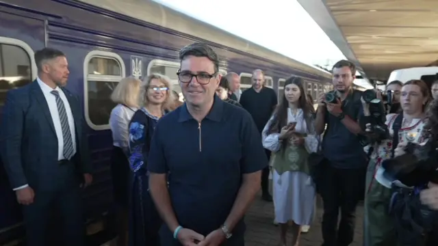 Any Burnham on a Ukrainian railway station platform, he is surrounded by several people.