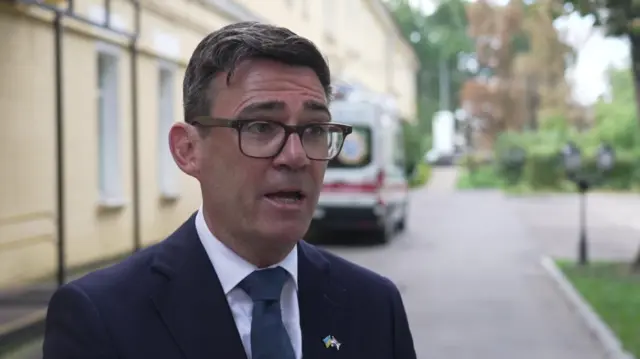 Andy speaks while wearing a navy suit and blue tie. He is wearing a badge of the UK and Ukrainian flags.