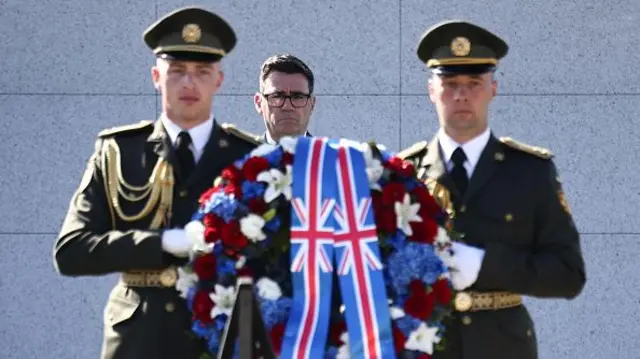 Andy Burnham stands behind two soldiers carrying a wreath during a visit to Ukraine. The wreath has two Union Flags on display