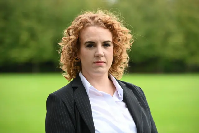 Cleveland police federation chairman Lauren Somerville. She has shoulder-length red curly hair and it wearing a white shirt and pinstripe black jacket. She is outdoors and the green grassy background is blurred
