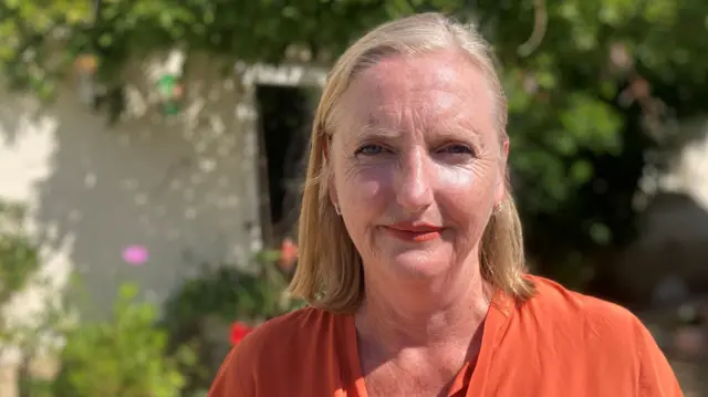 Melanie Kane has short straight blonde hair and wears an orange short-sleeved top with a bow. There is a tree and potted plants behind her.