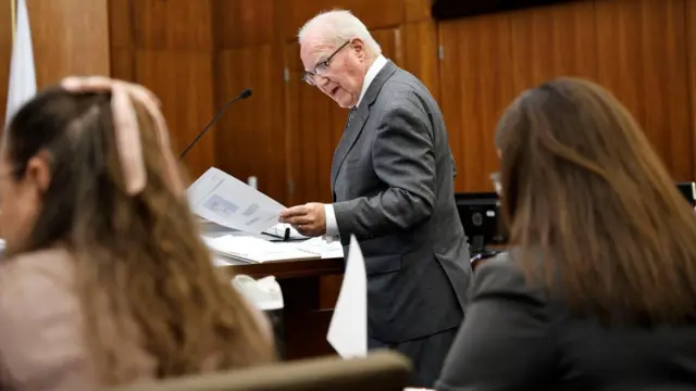 attorney, wearing a suit, holding papers and speaking into a microphone at a podium in a courtroom