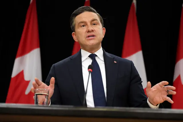 A man in a suit gestures from behind a lectern against a backdrop of Canadian flags
