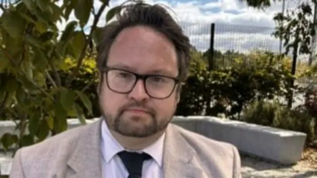Cleveland Police and Crime Commissioner Matt Storey is looking straight ahead and is wearing a beige suit jacket, white shirt and black tie. He has black rimmed glasses and a beard and moustache.