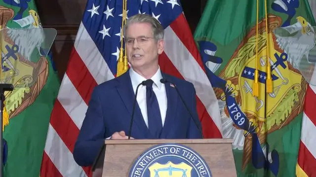 Scott Bessent standing at a podium in front of American and other colorful flags