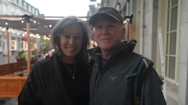 A man and a woman wearing rain gear smile at the camera, he has his arm around her