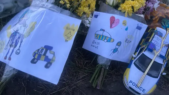 A yellow, blue and white toy police car is set among the floral tributes. There is a plastic flower set on top of it as well as a handwritten note. There are drawings of police officers and the word "RIP" on two pieces of paper wrapped around bouquets of flowers on the left of it.