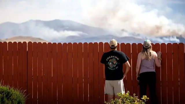 Two people look over what appears to be a domestic fence. They are looking at where smoke is rising from hills in the background.