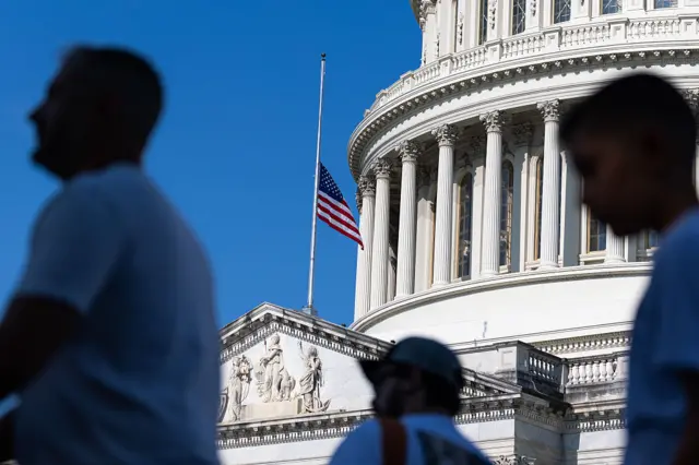 The U.S. flag is flown at half-staff over the U.S. Capitol Building, as people who have been blurred stand in the foreground.