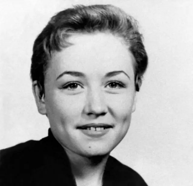 Dolly Parton, age 14, with short hair and a slight smile, in a black and white high school portrait., She wears a dark shirt and pendant necklace.