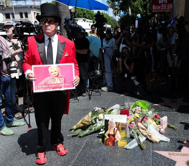 A man wearing a red tux holding a sign with Dolly's face and the words: We will always love you