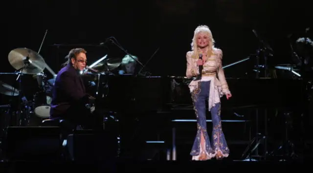 Elton John sits at a piano, while Parton stands and sings at a microphone