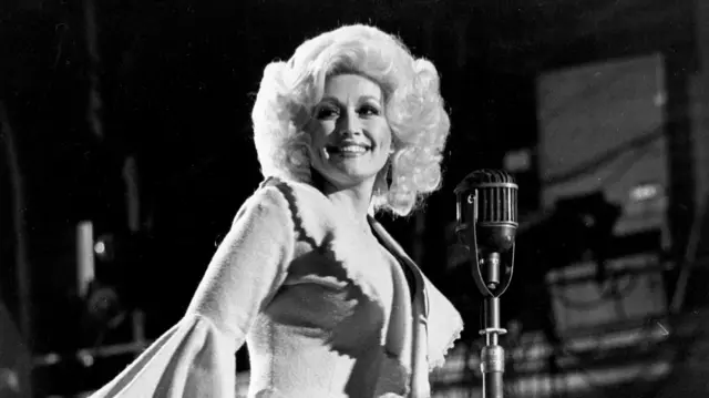 Dolly Parton photographed at the Grand Ole Opry. She wears a suit with bell sleeves and smiles as she stands by a microphone