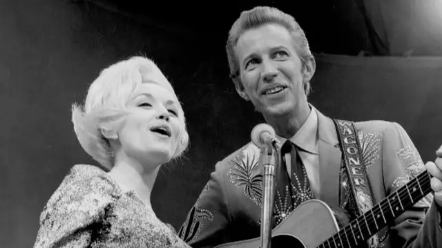 Dolly Parton has short blonde hair and Wagoner has a suit with diamantes on it