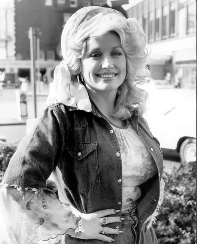 A black and white portrait of Dolly Parton wearing a head scarf and smiling while posing with her right hand on her hip