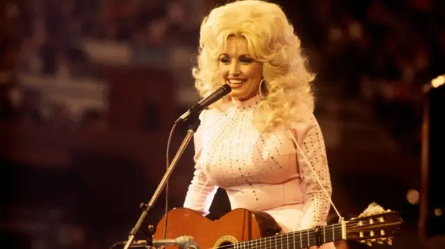Parton has big blonde hair and a pink shirt, she stands by a microphone with a guitar