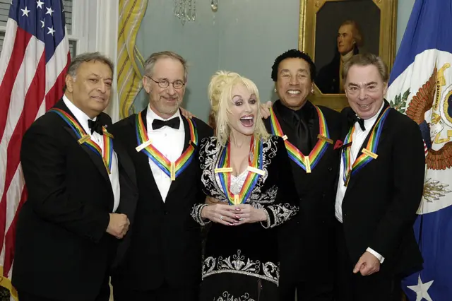 Four men and a woman wear dark suits and rainbow sashes and smile in front of a portrait and flags