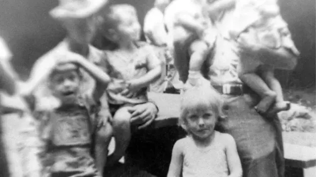 Dolly Parton, aged 5, with her family 1951 in a black and white image. She stands in a sleeveless white dress while adults holding children stand out of focus behind her.