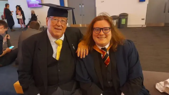 Matthew Wane with his father at his graduation ceremony. Matthew is on the right, wearing his graduation gown, while his father is sat on the left, with his arm on Matthew's shoulder while wearing Matthew's graduation hat