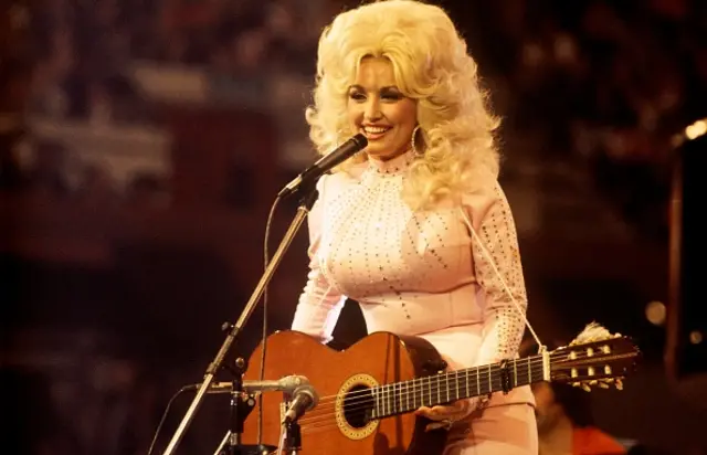 Dolly Parton smiles while holding a guitar in front of a microphone in 1976. She is wearing a white top with sequins on it