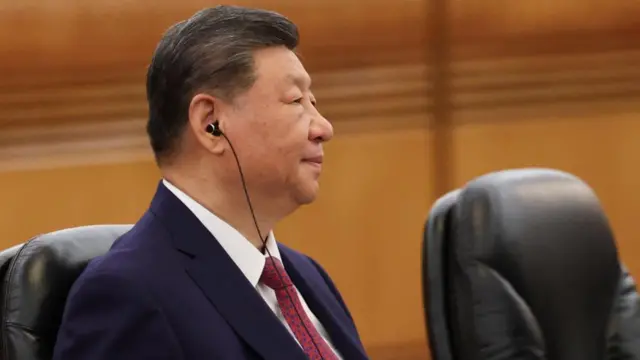 Xi Jinping is pictured sitting in a chair with a headpiece in his ear, wearing suit and tie.