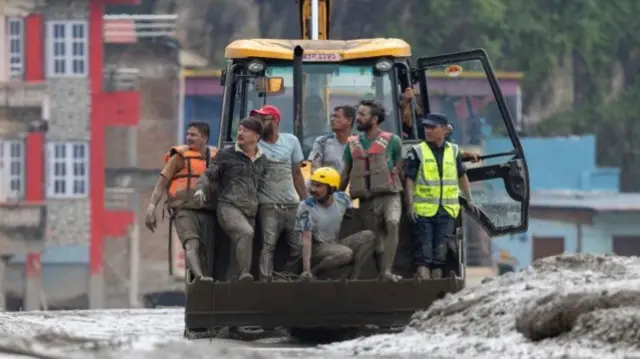 Seven men in muddy clothes are shown in the front scoop of a digger that is on a mud-covered street.