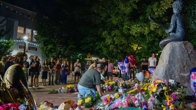 A fan crouches down at the base of a Dolly Parton statue, where flowers, balloons and cards have been left, as a crowd of people surrounds her