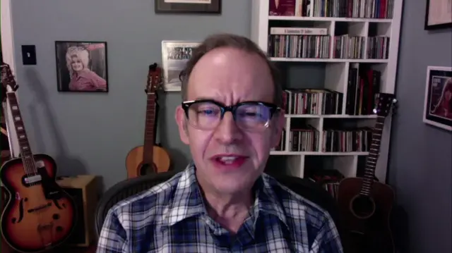 Bill DeMain is sat in what appears to be his, in front of a bookshelf filled with CDs and several guitars. There is also a picture of Dolly Parton on his wall