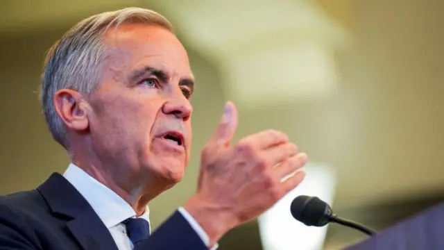 Mark Carney is pictured speaking behind a microphone, gesturing with his right hand.