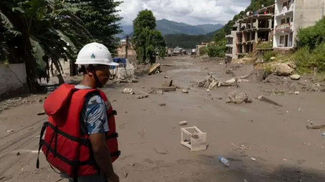 A man with a white hard hat and a red inflatable vest surveys a flooded landscape