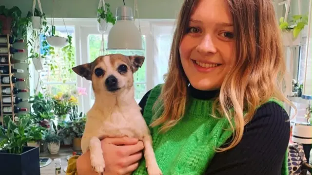Lili, 28, holds up Dolly the chihuahua. Both are looking at the camera. Lots of plants can be seen in the kitchen in the background